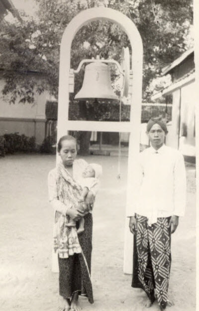 Vrouw met kind op de arm en man voor een vrijstaande klokkenstoel. Beiden gekleed in sarong.