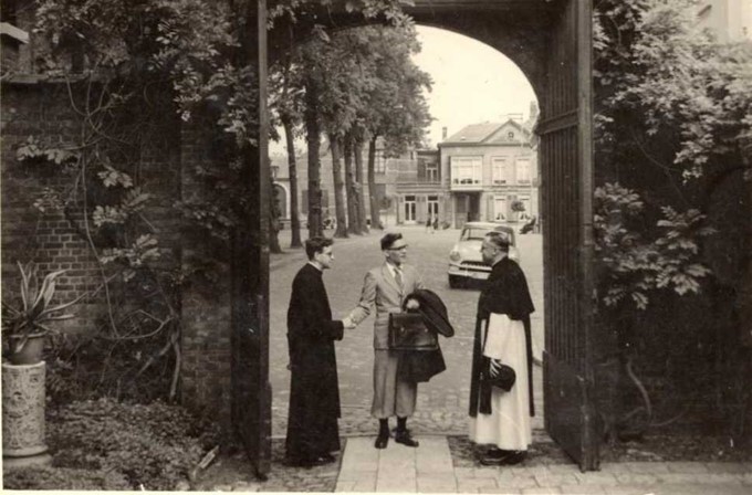 drie kruisheren, een in habijt, een in broederhabijt en een in reiskostuum staan in een geopende karrenpoort, een plein op de achtergrond