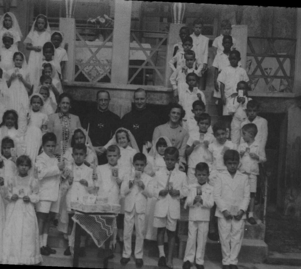 groep kinderen in witte communiekleding rond twee kruisheren in habijt voor een veranda