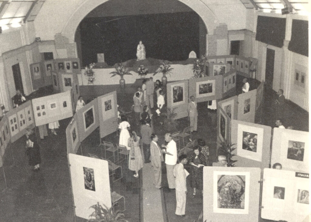 zicht vanaf hoogte op een zaal waarin mensen kijken naar reproducties van bekende maria-schilderijen die gemonteerd zijn op tentoonstellingsborden die in gebogen rijen door de ruimte staan
