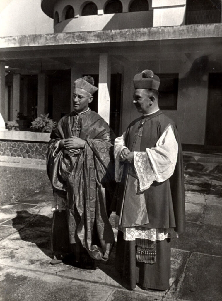twee monsignori ten voeten uit in de voor hun ambt officiële kledij staan in de zon voor een veranda