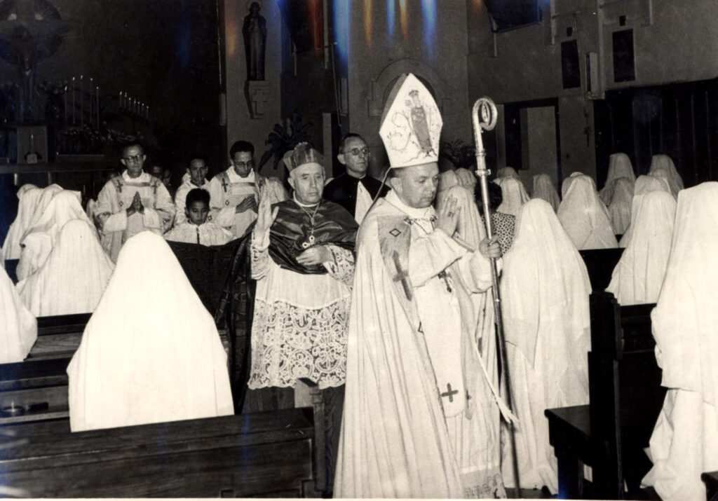 twee prelaten met gevolg trekken in plechtige processie zegenend de kerk uit tussen de banken met witgesluierde religieuzen door