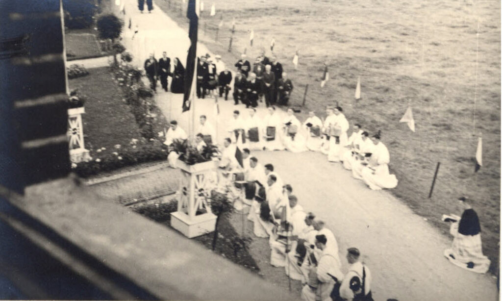 zicht vanaf hoogte op een groep kruisheren-diakens in liturgische kleding op een knie geknield op het versierde voorplein van het klooster