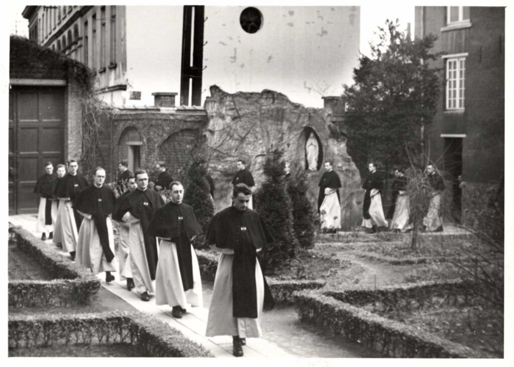 de kruisheren in Diest lopen achter elkaar over een tegelpaadje vanuit de kerk door de kloostertuin naar de kloostergang, op de achtergrond de grote poort en de Lourdesgrot